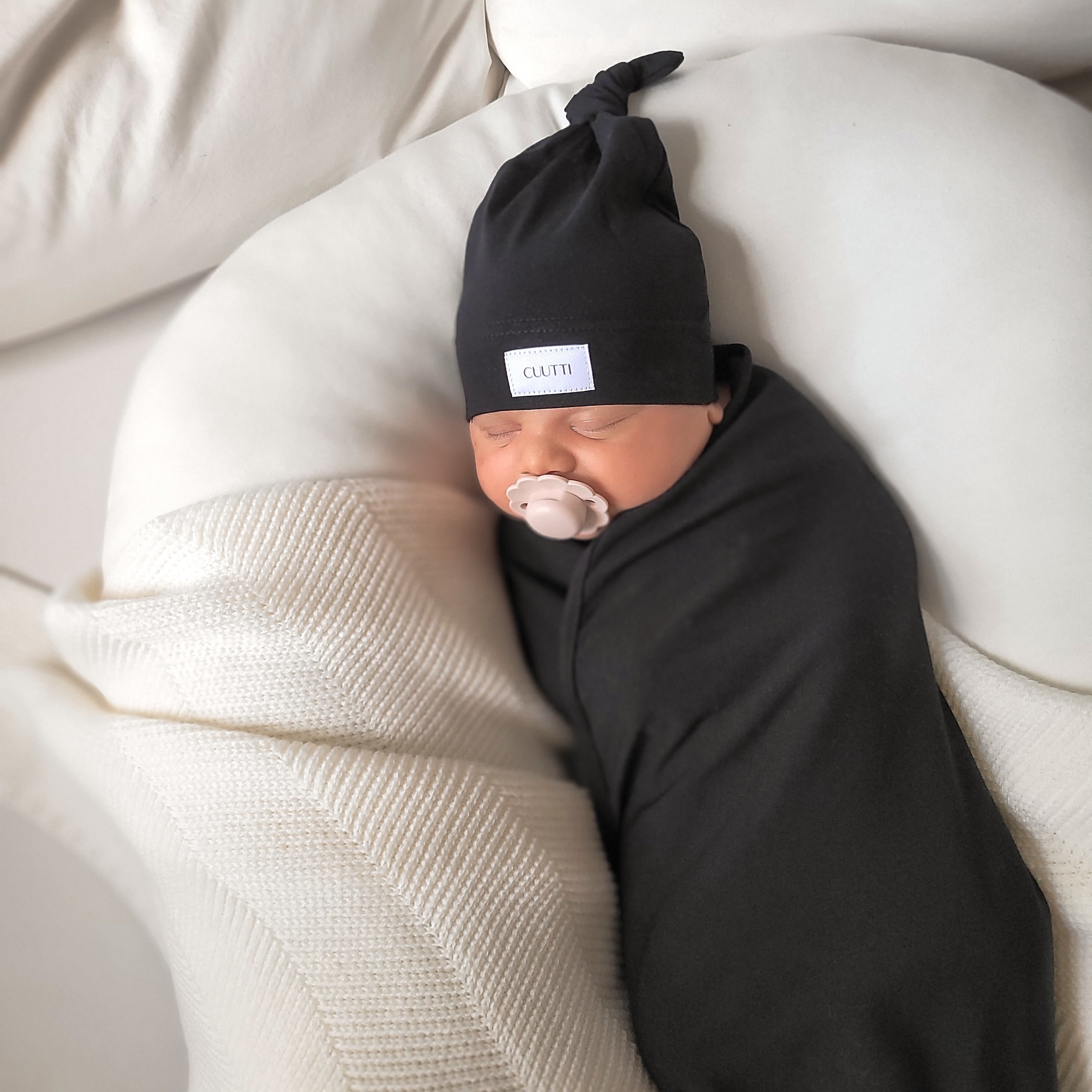 A lovely newborn baby boy sleeps soundly with a pacifier, cozy in a black CUUTTI baby swaddle and a matching black CUUTTI beanie.