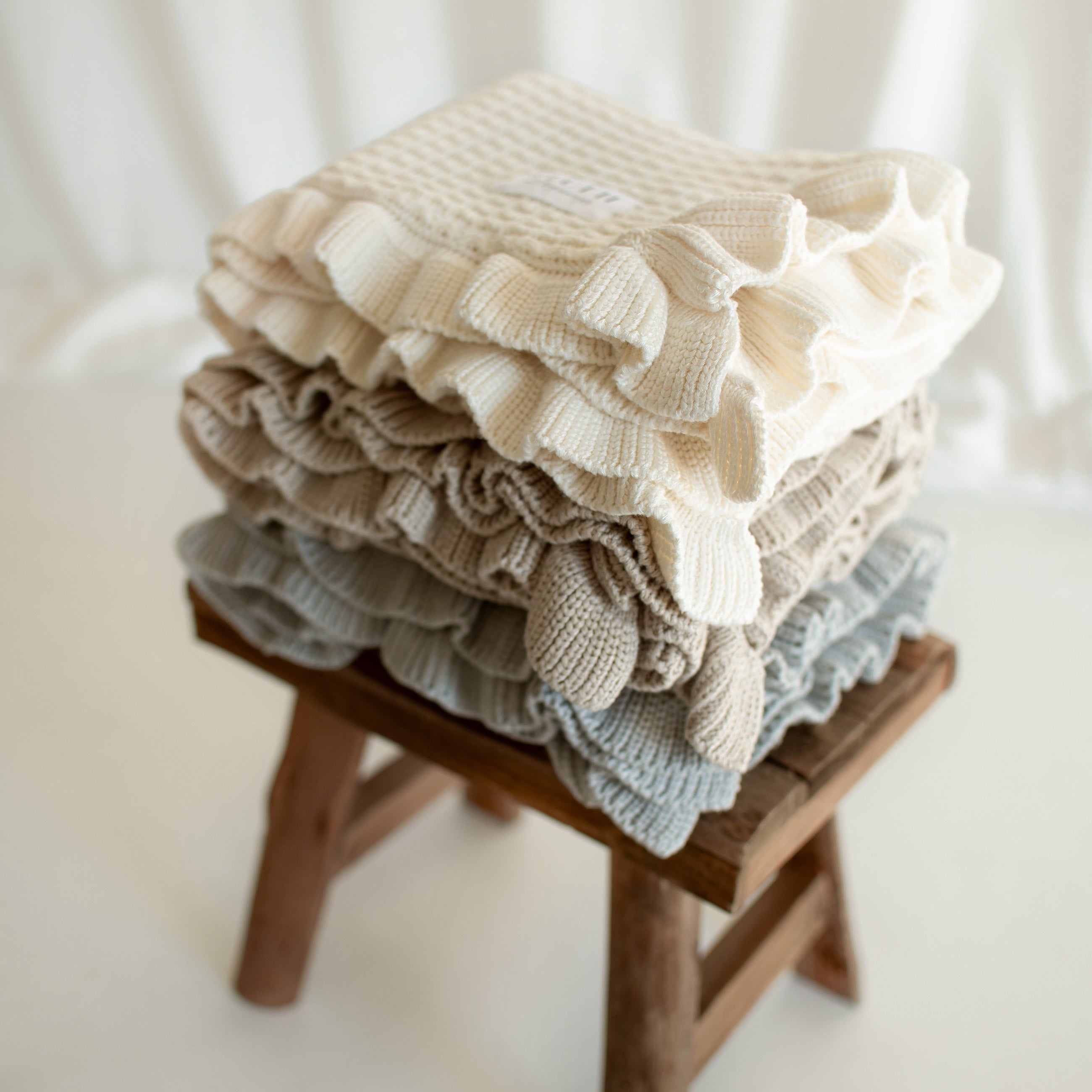 Three CUUTTI Ballerina bamboo blankets in ivory, beige, and cloud color neatly folded on a small wooden bench.