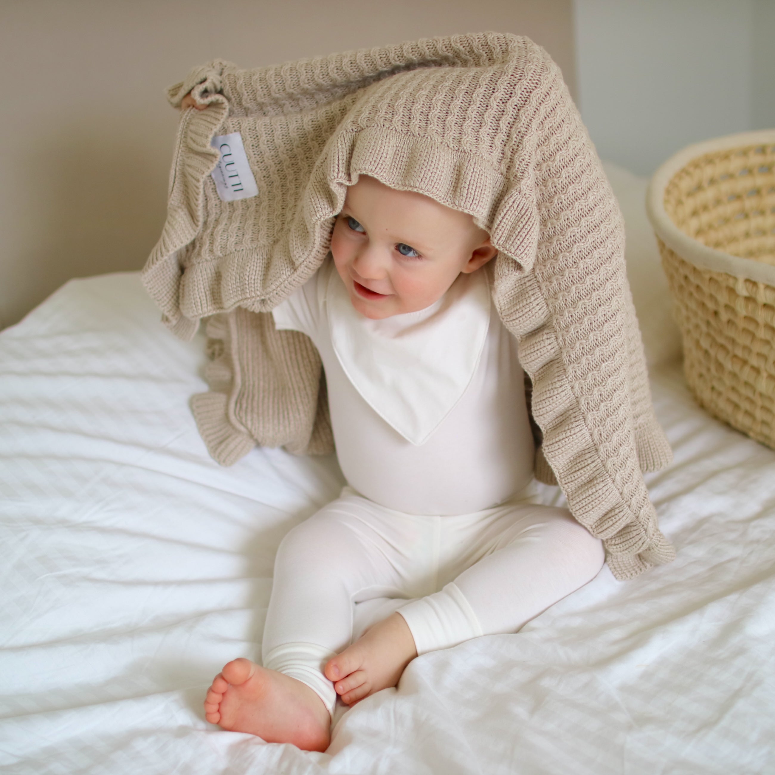 Little boy on a bed, playfully hiding under a beige CUUTTI Ballerina blanket to tease his parents.