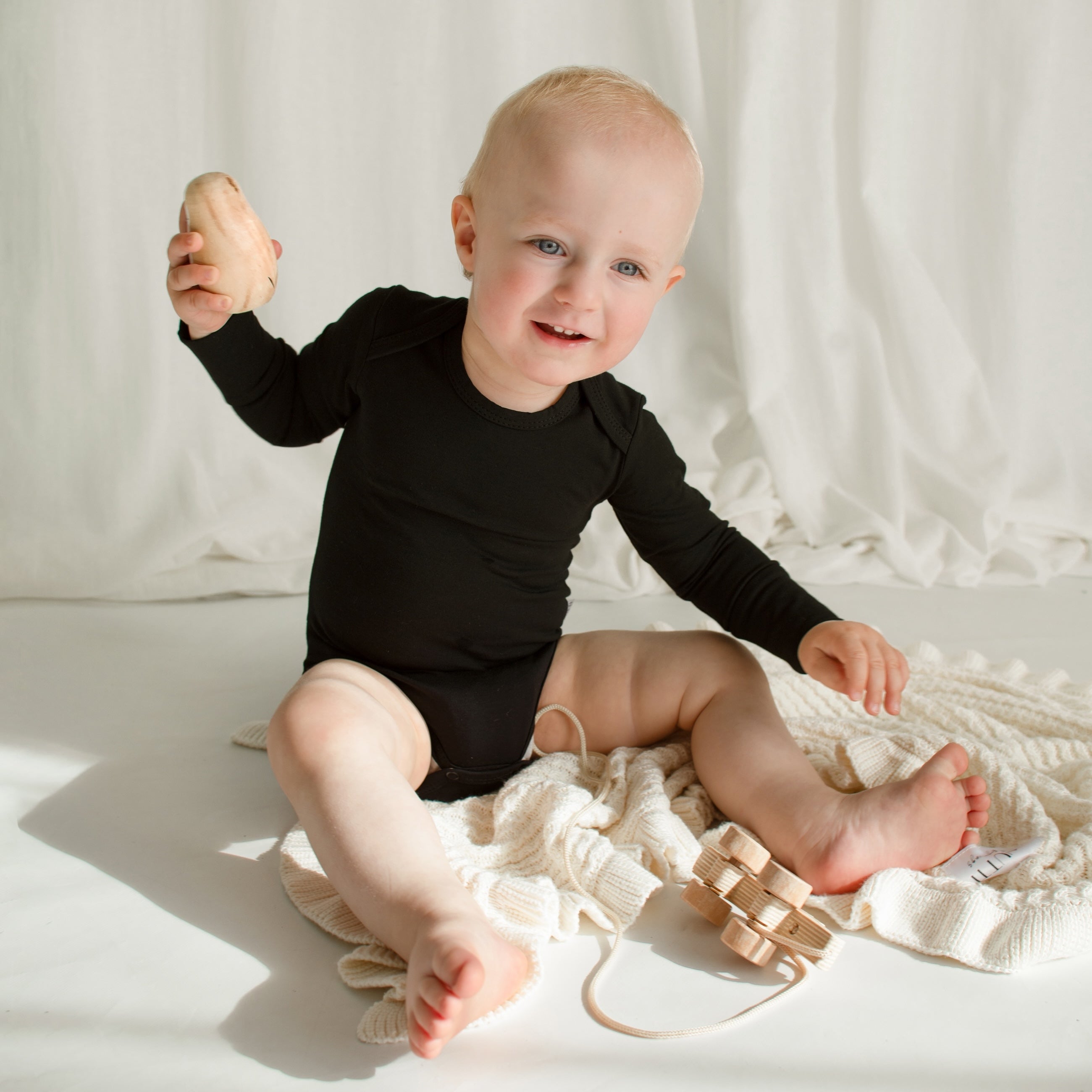 Little boy happily playing on the floor atop a CUUTTI Ballerina bamboo blanket in ivory, dressed in a black CUUTTI long sleeve bodysuit.