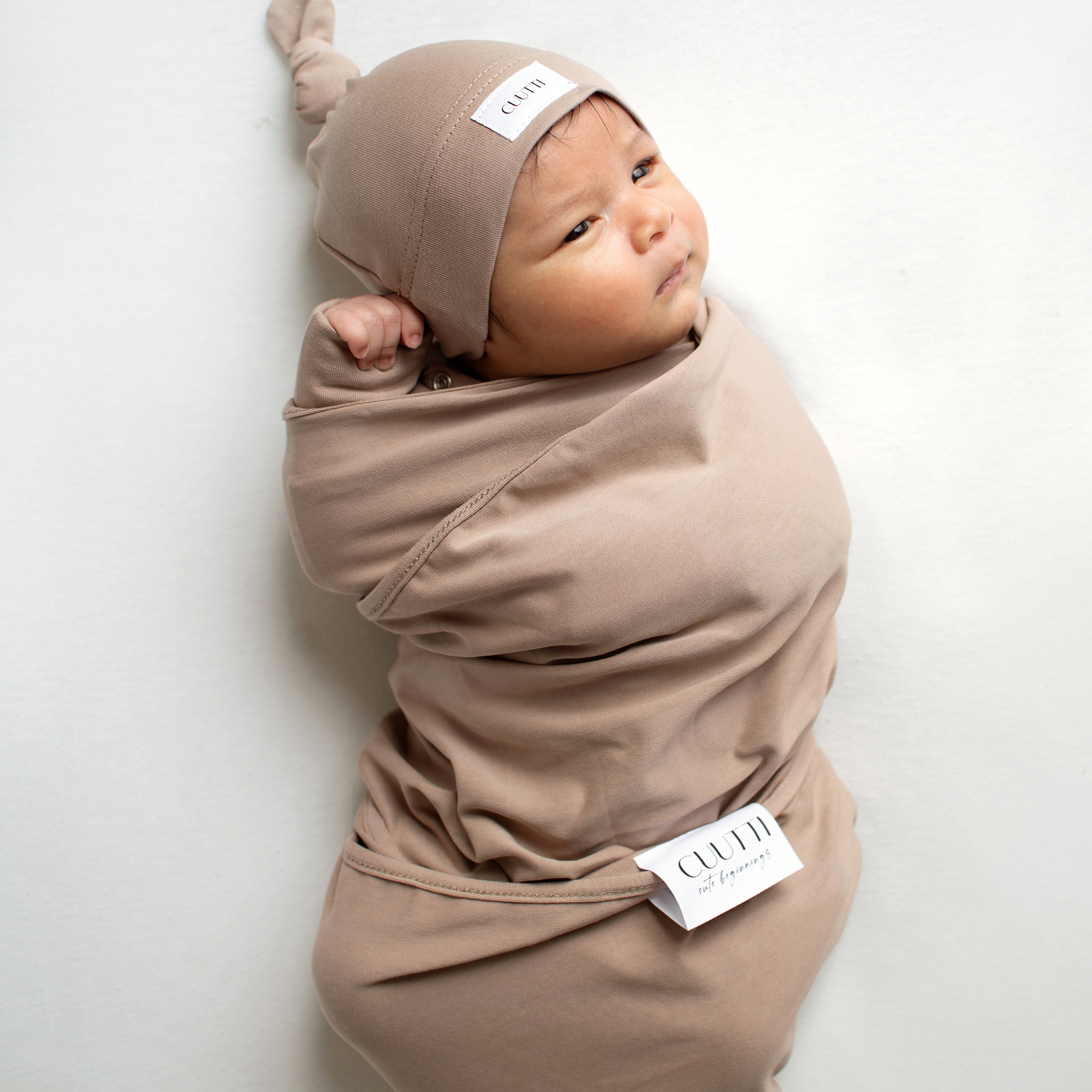 A 1-month-old baby boy wrapped in a CUUTTI cookie-colored baby swaddle and wearing a matching CUUTTI cookie-colored baby beanie.