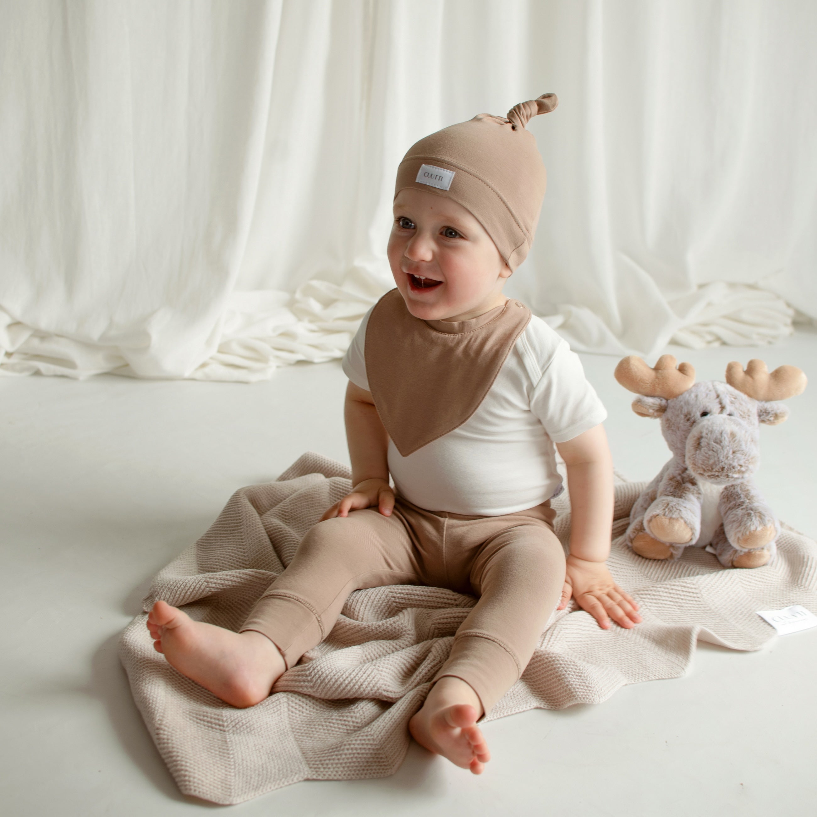 Adorable baby boy sitting on the floor wearing a CUUTTI baby beanie, waterproof bib, and leggings, all in cookie color, with an ivory short-sleeve bodysuit.