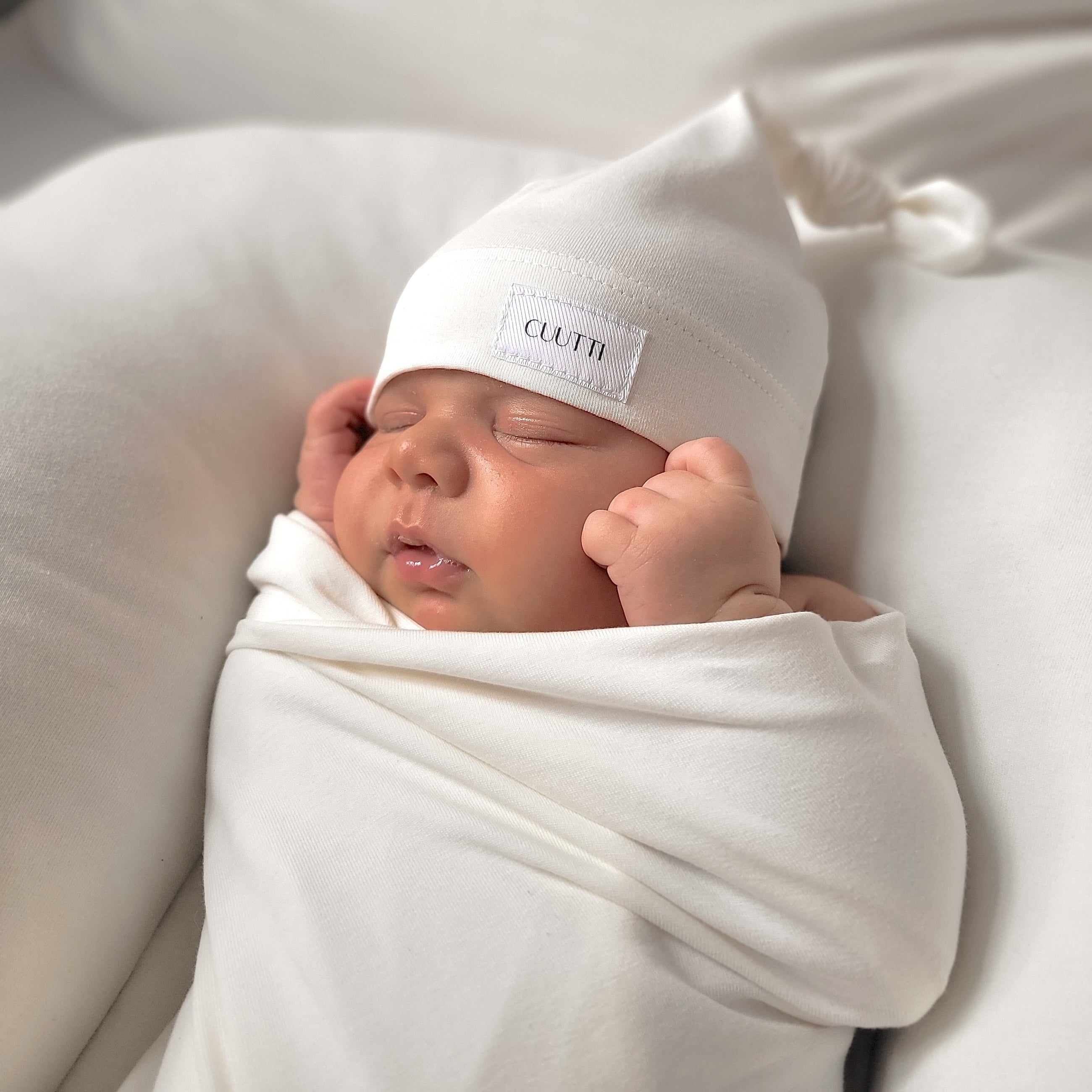 Close-up of a newborn baby boy sleeping peacefully, wrapped in an ivory CUUTTI baby swaddle and wearing a matching ivory CUUTTI baby beanie.