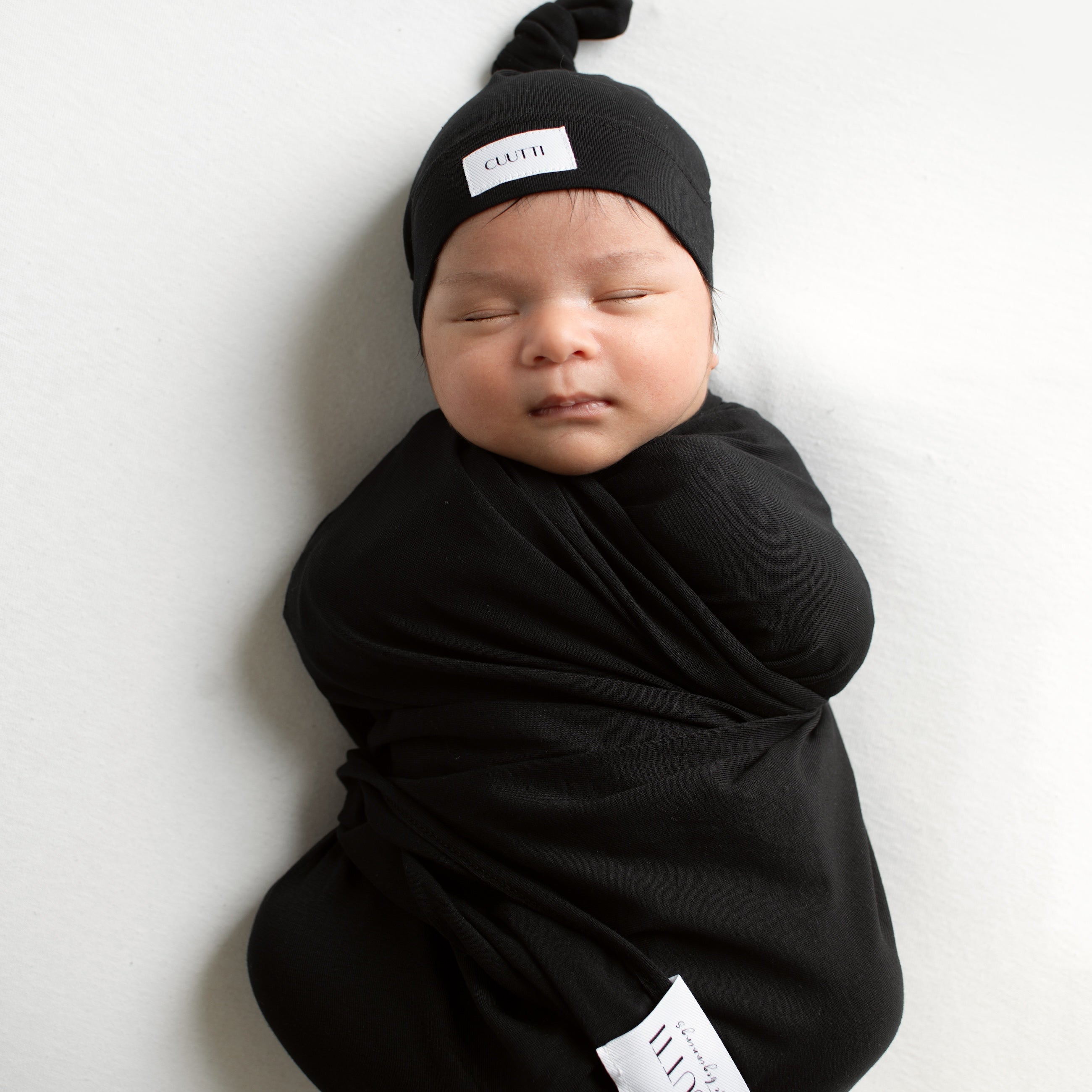 1-month-old baby boy wrapped in a black CUUTTI baby swaddle and wearing a matching black CUUTTI baby beanie.