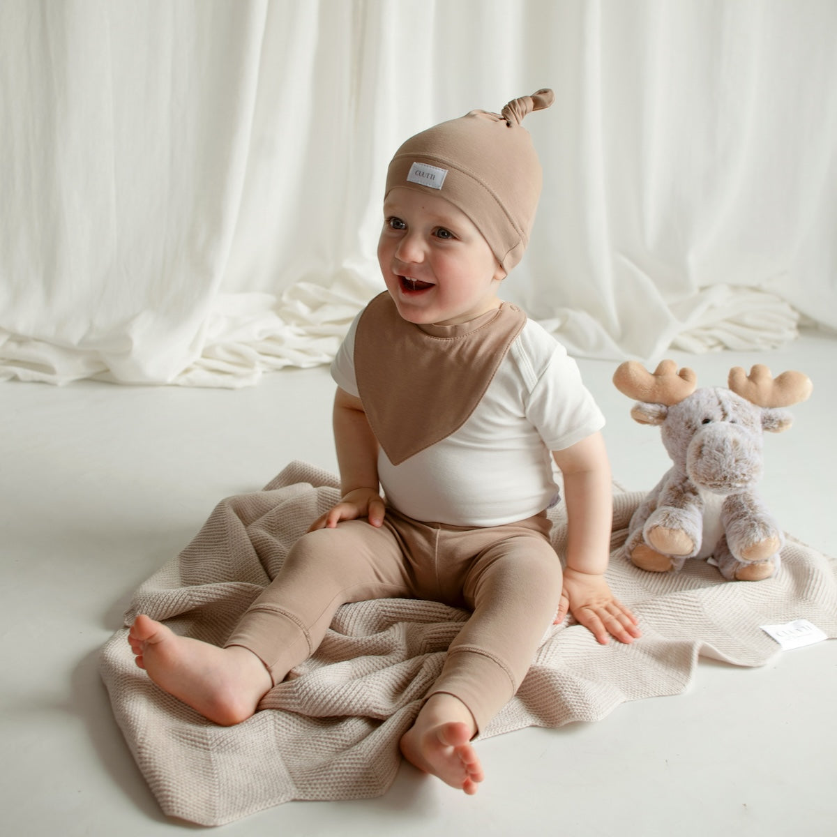 Adorable baby boy sitting on the floor wearing a CUUTTI baby beanie, waterproof bib, and leggings, all in cookie color, with an ivory short-sleeve bodysuit.