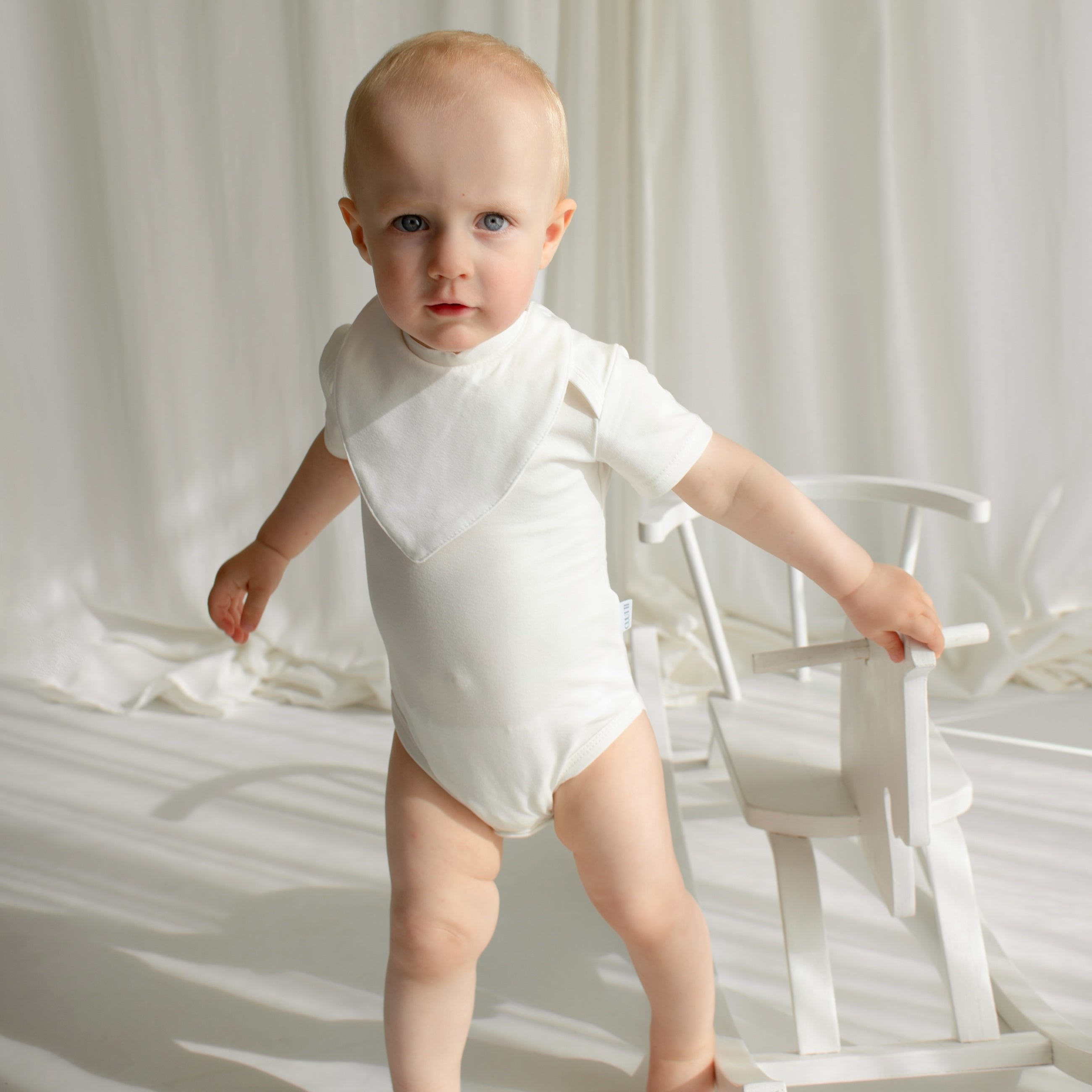 Little boy standing next to a rocking horse, wearing an ivory CUUTTI short sleeve bodysuit and a matching ivory CUUTTI waterproof bib.