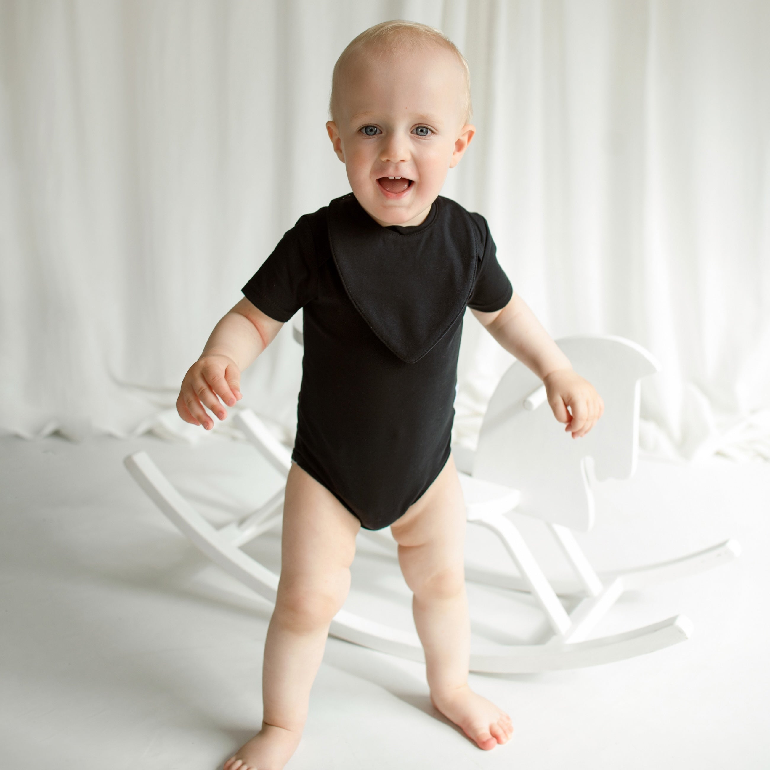 Proud little boy standing and highlighting his stylish CUUTTI look, including a black short sleeve bodysuit and a matching black waterproof bib.