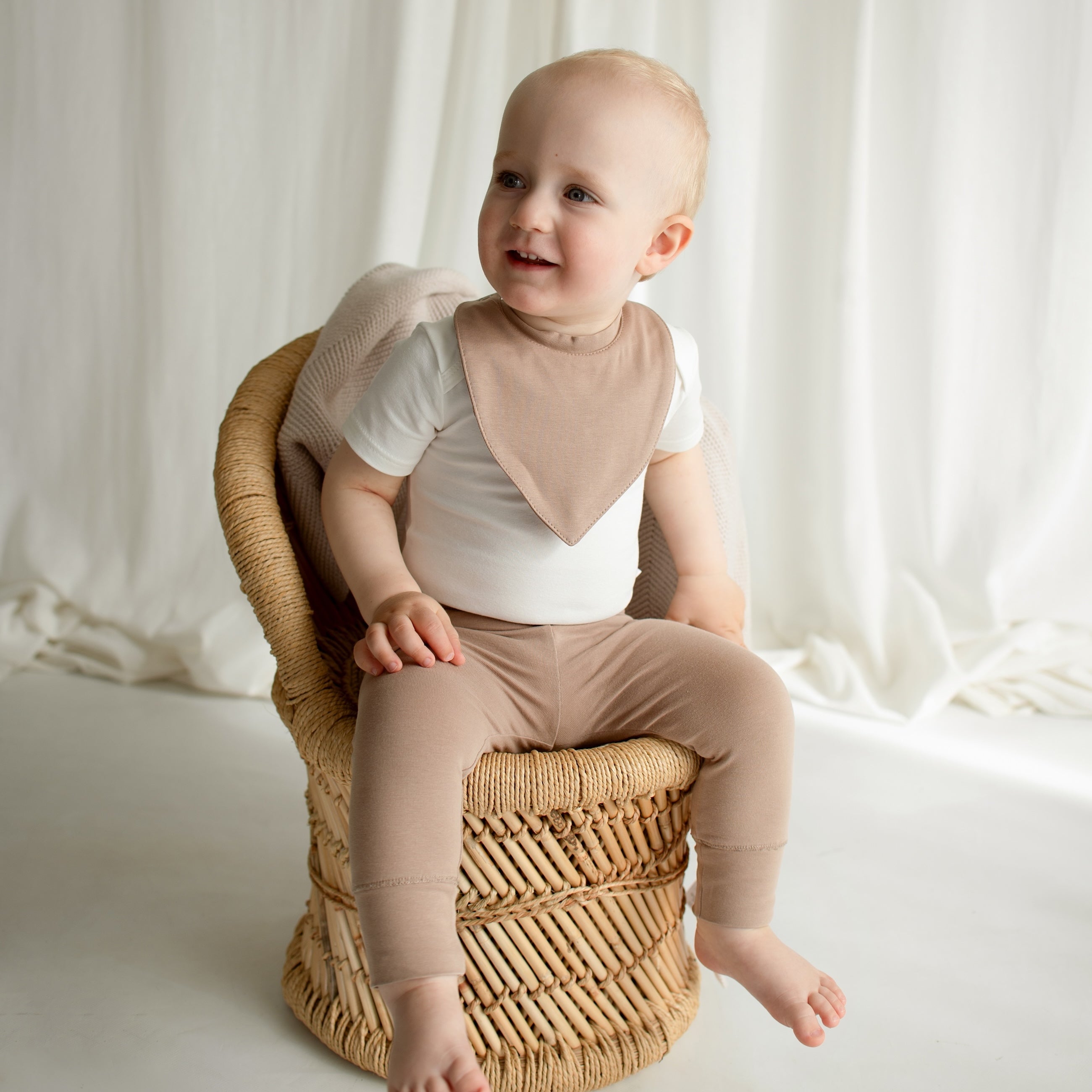 A little boy sitting on a small rattan chair, wearing a stylish CUUTTI outfit featuring an ivory short sleeve bodysuit, a cookie-colored waterproof bib, and cookie-colored leggings.