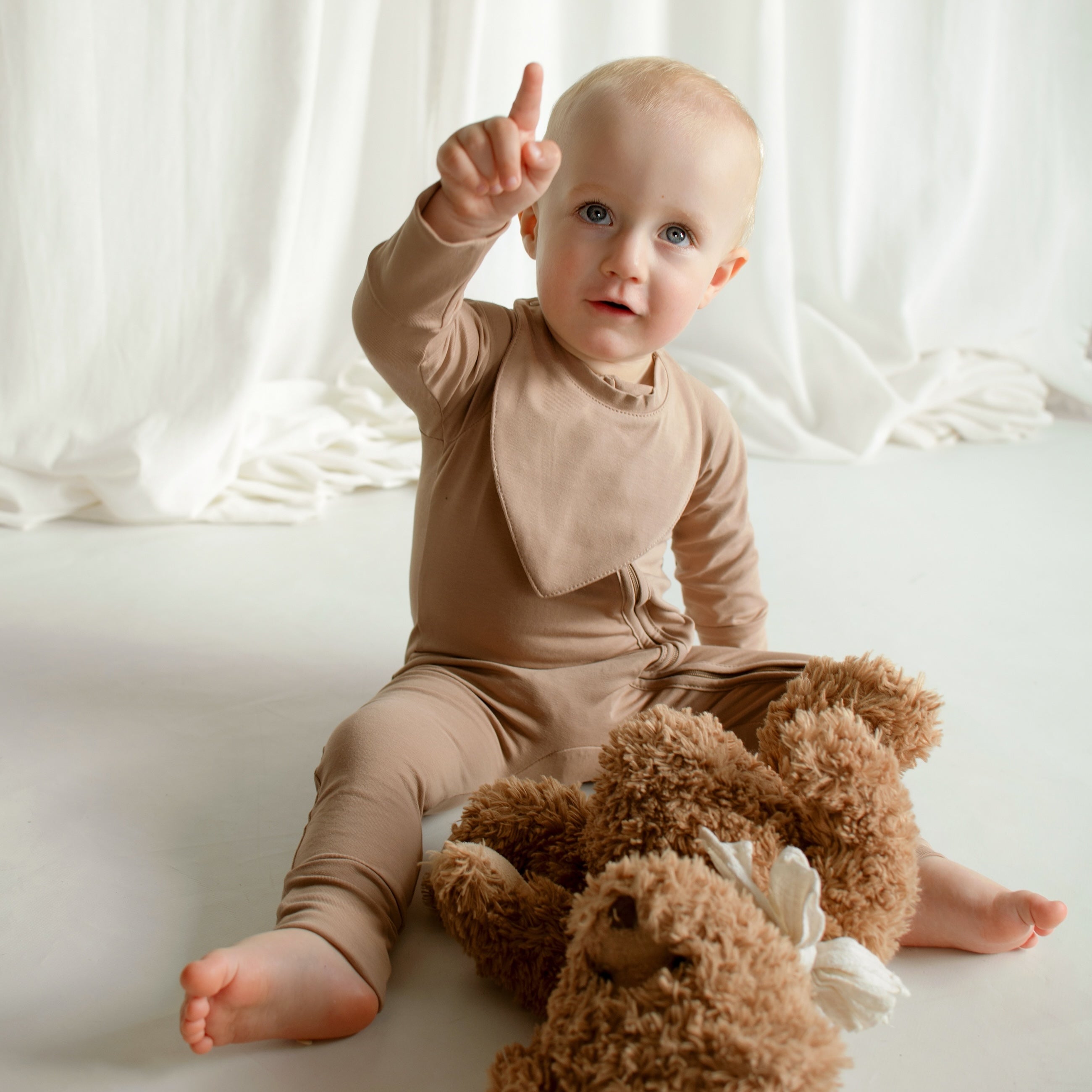 A little boy sitting on the floor in a cookie-colored CUUTTI zipper onesie and a matching cookie-colored CUUTTI waterproof bib.