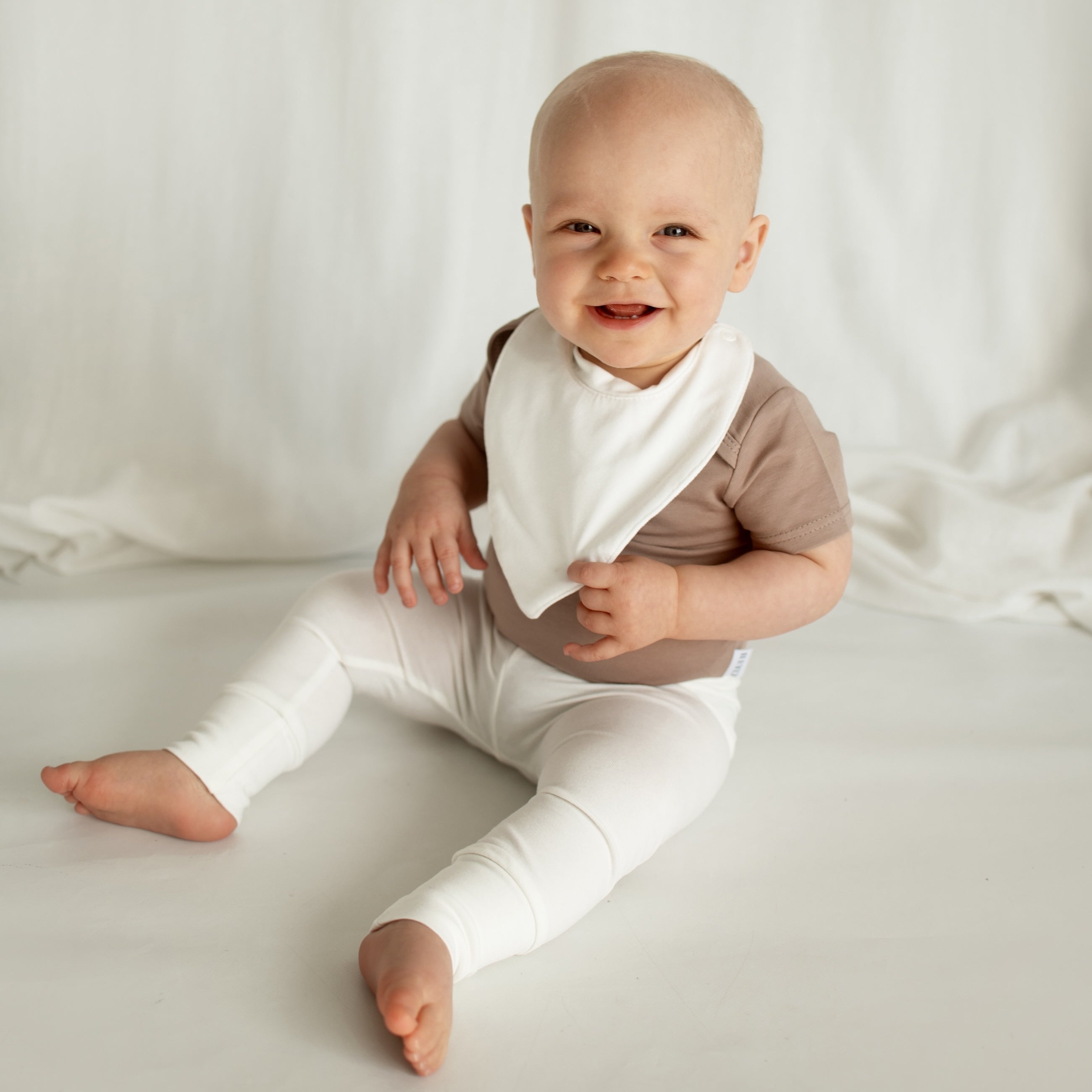 A smiling little girl sitting on the floor in a stylish CUUTTI outfit, featuring a cookie-colored short sleeve bodysuit, ivory waterproof bib, and ivory leggings.