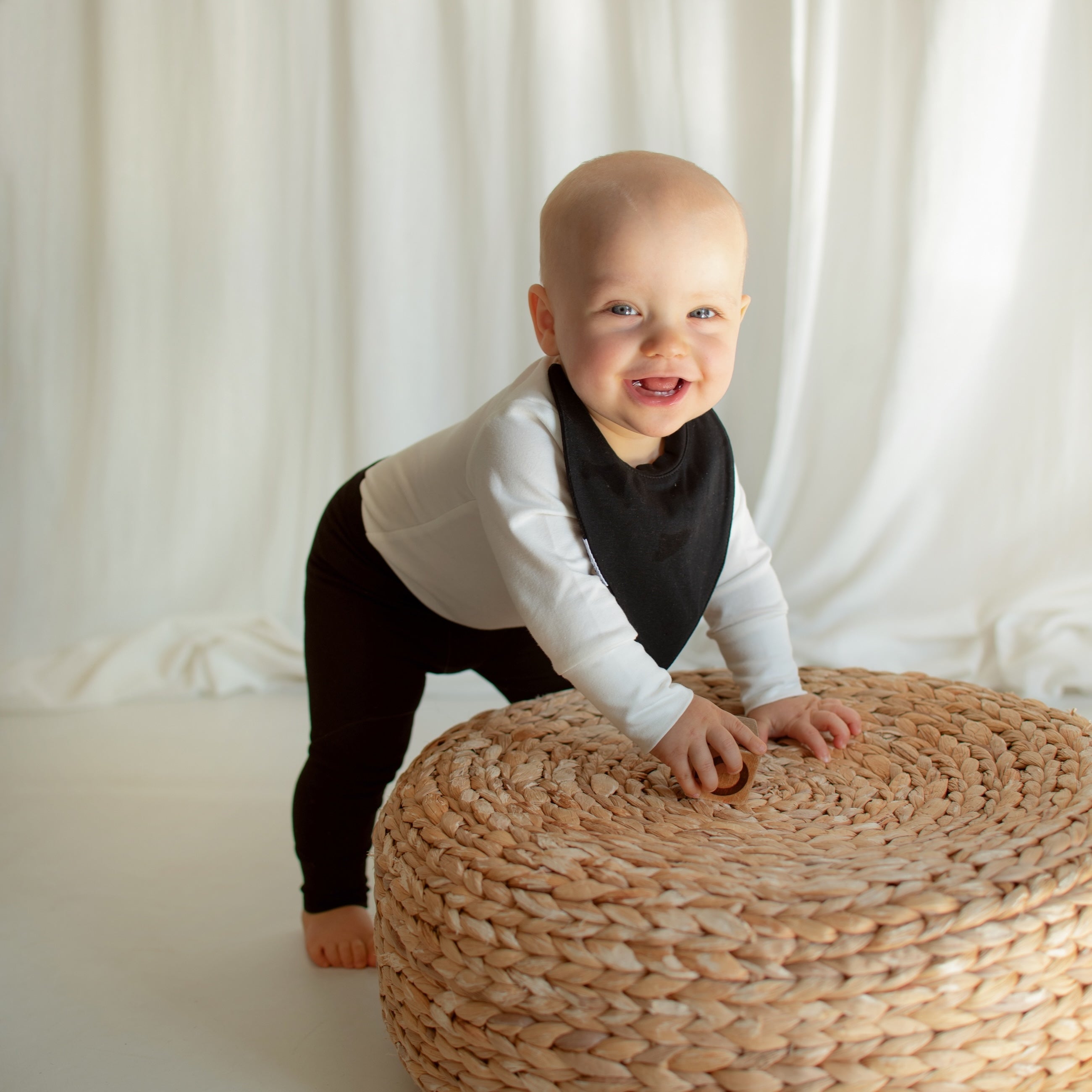 Smiling little girl practicing standing against a rattan footstool in a stylish CUUTTI outfit, featuring a white long sleeve bodysuit, black waterproof bib, and black leggings.